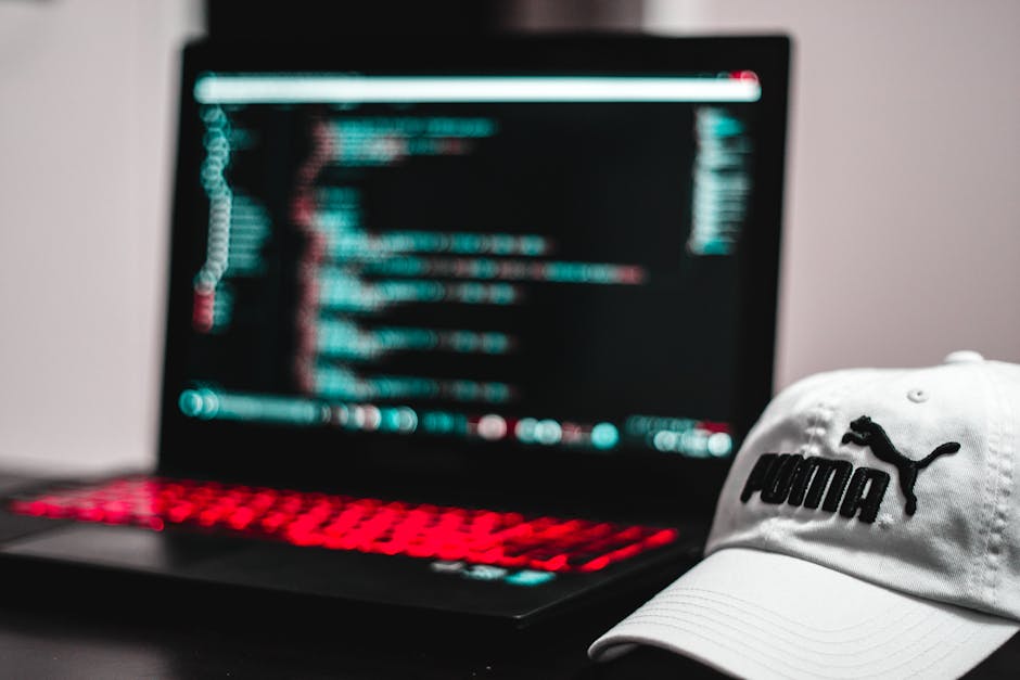 A black laptop with a red keyboard illuminated by red backlighting, displaying a screen filled with lines of code in bright colors such as blue, red, and white, placed on a flat surface. Next to the l